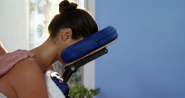 A woman is receiving a professional shoulder massage while sitting on a padded chair. The therapist's hand is gently placed on her shoulder. This image can be used for promoting wellness, self-care tips, massage therapy services, relaxation techniques, and health and spa treatments.