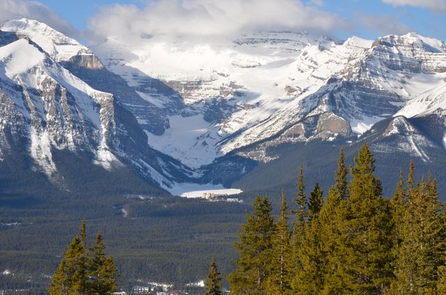 Winter scene with snow-covered peaks of Rocky Mountains and dense pine forest in foreground. Rocky cliffs contrast with green trees below, creating a picturesque and serene landscape. Ideal for themes like outdoor adventures, nature photography, travel destinations, and wilderness exploration.