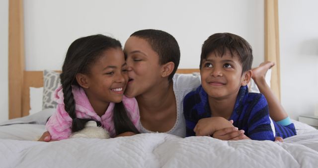 Mother and two children lying on bed, mother kissing daughter, son smiling. Perfect for family-themed marketing, parenting blogs, advertisements focused on familial love and togetherness, or articles promoting quality time at home.