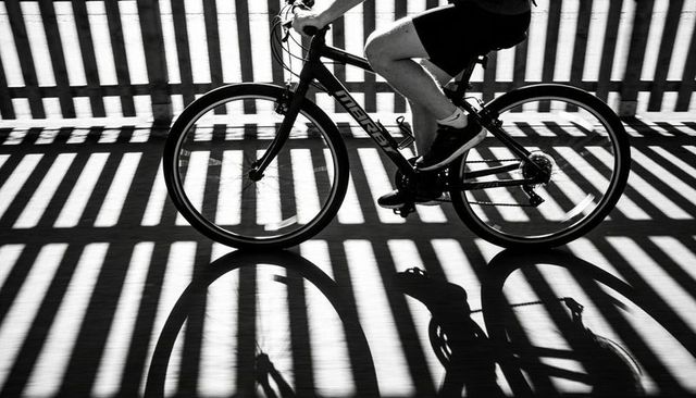 Cyclist pedaling through dramatic striped shadows on sunlit pavement