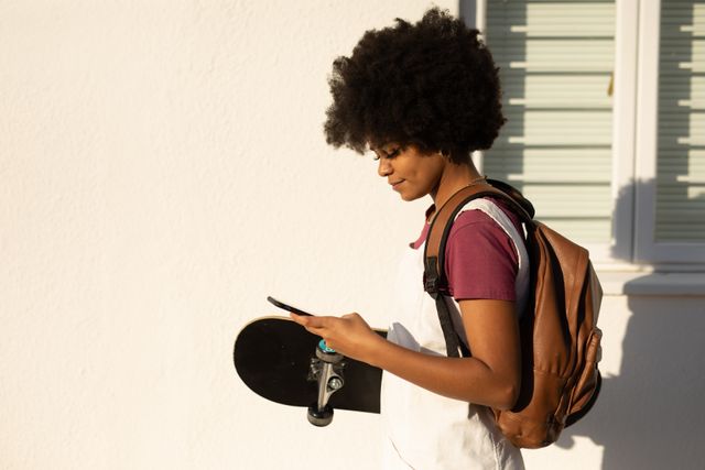 Young woman with afro hair walking on a sunny day, carrying a skateboard and using a smartphone. Ideal for themes related to urban lifestyle, youth culture, technology, communication, and leisure activities.