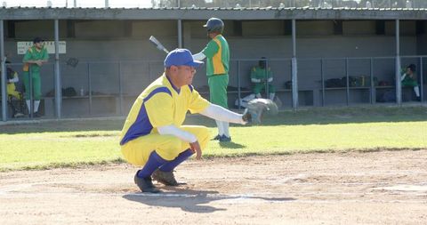 Veteran catcher squatting at plate wearing yellow-blue uniform with batter in green dugout