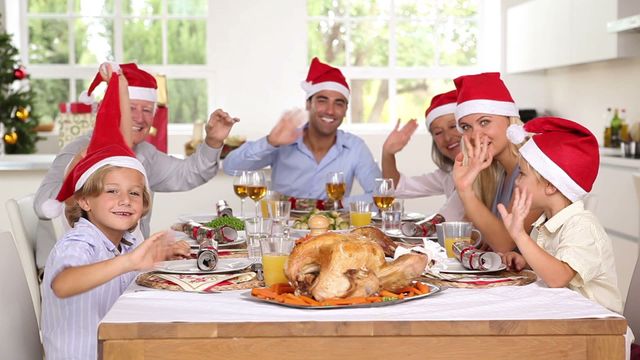 Family members of multiple generations are gathered around a table, smiling and waving while wearing Santa hats. They enjoy a festive holiday meal that includes a beautifully prepared turkey with various side dishes. The scene is cheerful and filled with joy, making it suitable for use in advertisements, Christmas greeting cards, holiday promotional materials, or any context that emphasizes family gatherings, holiday festivities, and joyful celebrations.