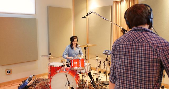 Woman playing drums while man adjusts sound engineering equipment, capturing dynamic recording session in a well-equipped professional studio. Suitable for illustrating music production processes, studio work, or educational content on sound engineering.