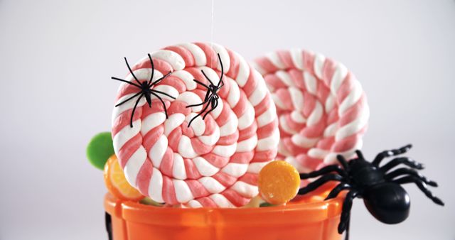 Halloween candy spirals are adorned with plastic spiders, creating a festive and spooky display in an orange bucket. The arrangement captures the playful and eerie spirit of Halloween celebrations.