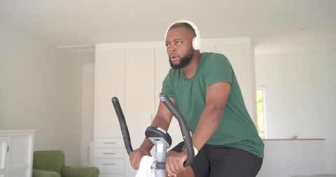 African american man pedaling on stationary bike at home wearing headphones and smartwatch