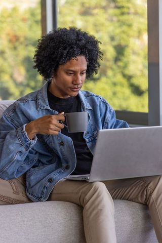 African american man working on laptop while drinking coffee on sofa in denim jacket