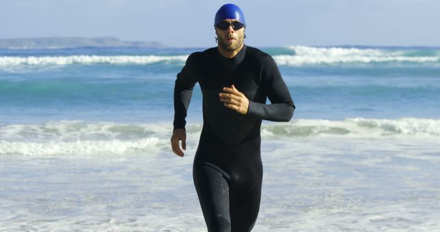 Man in wetsuit and goggles running from ocean to beach. Suitable for athletic, training, and water sports scenarios. Can be used to illustrate themes of fitness, perseverance, and outdoor activity.