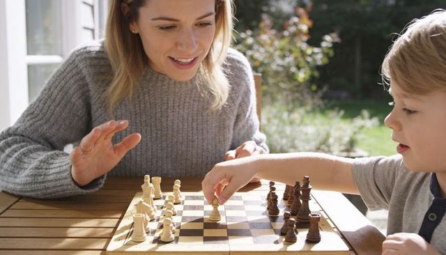 Young woman teaching child chess on sunlit backyard patio, playing wooden chessboard game
