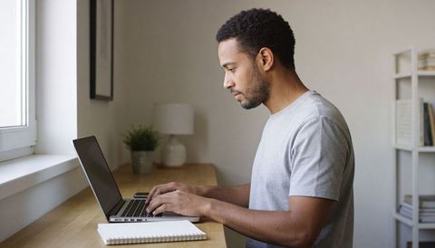 Man typing at window counter working on laptop in minimalist home office natural light