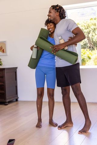 African american couple hugging and smiling after home yoga, holding mats and water bottles