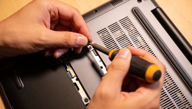 Hands tightening small phillips screw on laptop underside with black-yellow screwdriver