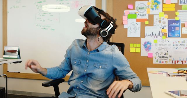 This photo captures a male designer wearing a VR headset, fully immersed in a virtual reality experience in a creative office setting. The surrounding environment includes whiteboards with brainstorming notes, project plans, and pinned graphics, indicating a collaborative and innovative workspace. This image is ideal for illustrating themes of modern technology, immersive experiences, innovation, creative work environments, and tech-savvy professionals. It can be used in articles, blogs, or promotional materials related to virtual reality, tech startups, modern workspaces, and creative industries.