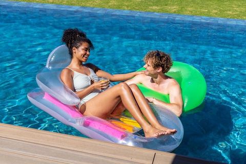 Diverse couple relaxing on colorful pool floats enjoying sunny backyard summer drink