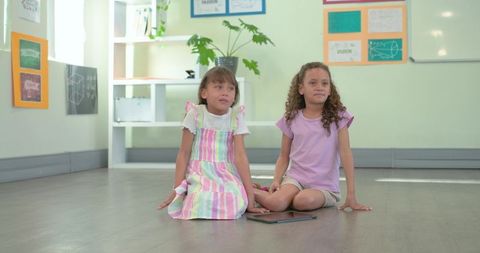 Two african american girls sitting on classroom floor sharing tablet and learning together