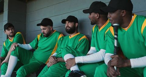 Sitting baseball team resting on dugout bench wearing green and yellow uniforms and black caps