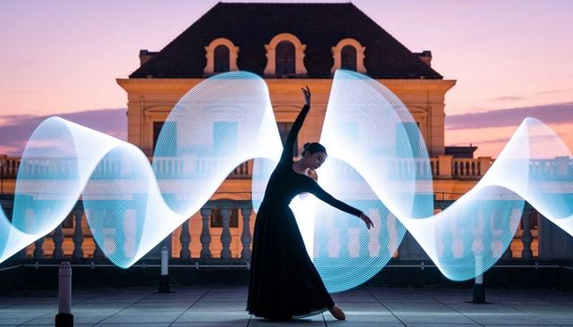 Ballerina performing en-pointe with neon ribbon light trails on rooftop terrace at dusk