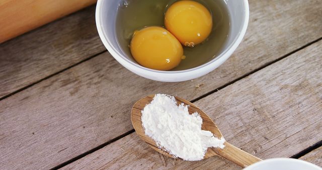 Raw baking ingredients on a rustic wooden table showcase two raw eggs in a bowl and flour on a wooden spoon, highlighting a step in the baking process. Ideal for illustrating culinary content, recipe ideas, cooking blogs, and kitchen-related articles.