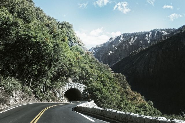 Depicts winding road through forest leading into tunnel with mountains in background. Ideal for themes of travel, adventure, nature, or road trips. Can be used in tourism, transportation services, or outdoor adventure promotions.