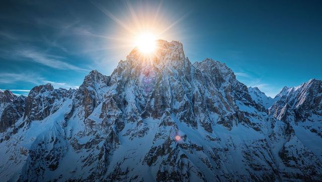 Sunburst crowning rugged alpine peak with snow-draped ridges and blue winter sky