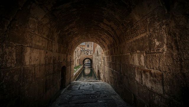 Barrel-vaulted stone tunnel framing narrow canal with cracked paving, arches and reflections