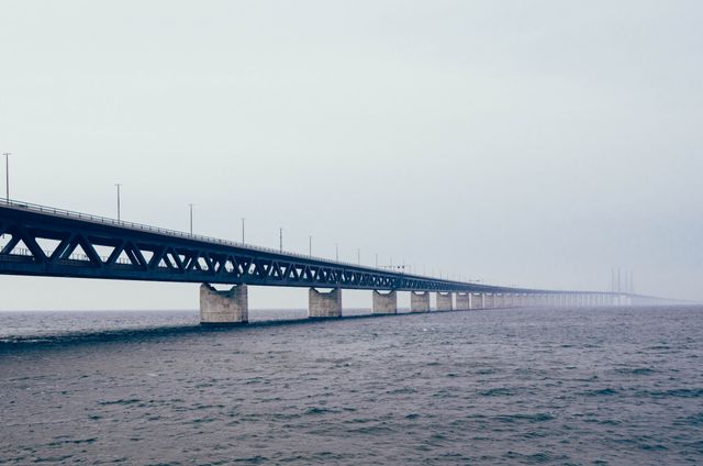 This image shows a long bridge extending over a calm sea under misty weather conditions. The fog creates a tranquil and mysterious atmosphere. Use for concepts of travel, journeys, transportation infrastructure, engineering, and calming scenic views. Ideal for travel brochures, engineering presentations, and websites focusing on journey and adventure themes.