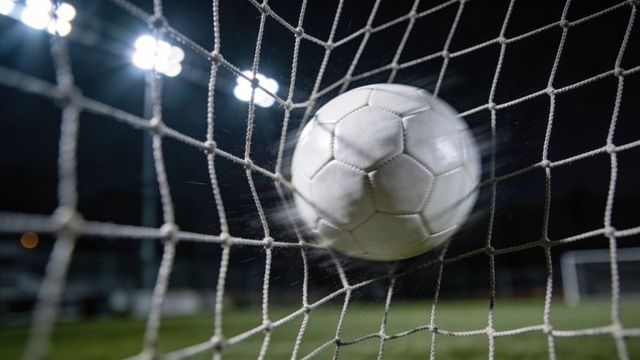 Striking soccer ball bulging goal net under stadium floodlights capturing goal impact