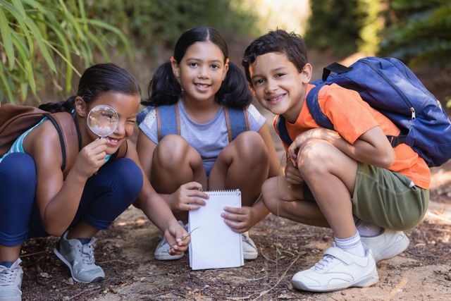 Three children are exploring nature in a park. One child is holding a magnifying glass, another is holding a notebook, and all are smiling. They are dressed casually and appear to be on an outdoor adventure. This image is perfect for educational materials, advertisements for outdoor activities, or promoting teamwork and curiosity among children.