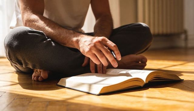 Man reading and annotating open book on sunlit parquet floor holding pen, morning light