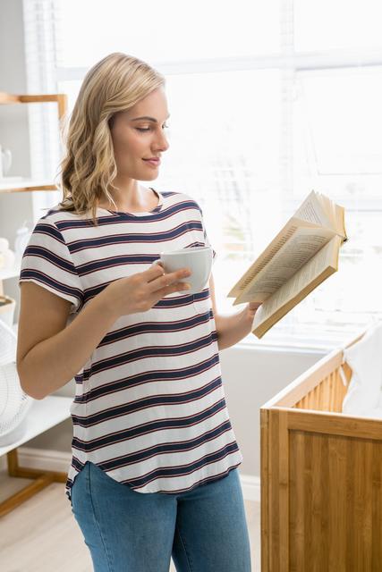 A woman with blonde hair, wearing a striped shirt and blue jeans, stands in a home interior while holding a coffee cup and reading a book. The sunlight streams through the window, creating a warm and cozy atmosphere. This image is ideal for lifestyle blogs, advertisements for home goods, or articles about relaxation and self-care.
