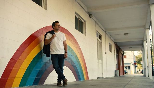 Man leaning against rainbow mural wearing casual t-shirt holding jacket and wristwatch