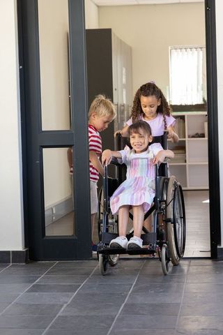 Multiracial children pushing smiling girl in wheelchair through school doorway