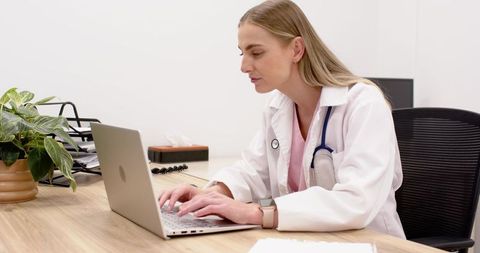 Female doctor typing on laptop in modern clinic office with stethoscope and green plant