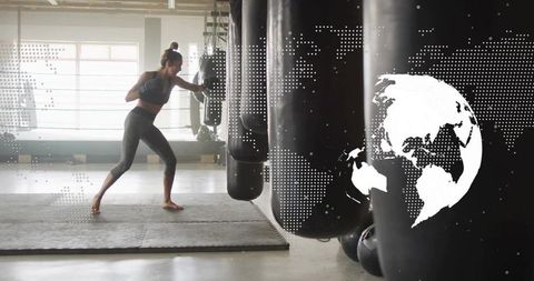 Female athlete punching heavy bag during boxing training in modern daylight gym, barefoot