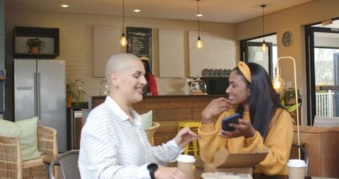 Two women laughing and sharing phone in cozy cafe with takeaway coffee and pastries