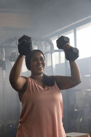 Confident african american woman pressing dumbbells overhead smiling during gym workout