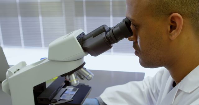 Scientist in lab coat observing a specimen through a microscope in a laboratory setting. Ideal for use in articles or presentations about scientific research, biology experiments, laboratory work, innovations in science, or educational materials emphasizing the importance of scientific discovery.