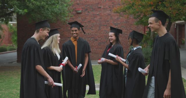 Front view of multi-ethnic group of high school teenage students wearing caps and gowns, holding diplomas and talking excitedly on their graduation day, laughing and smiling, taking off their caps and throwing them in the air in celebration 
