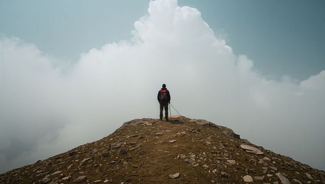 Hiker standing on mountain ridge facing towering clouds with red backpack and poles