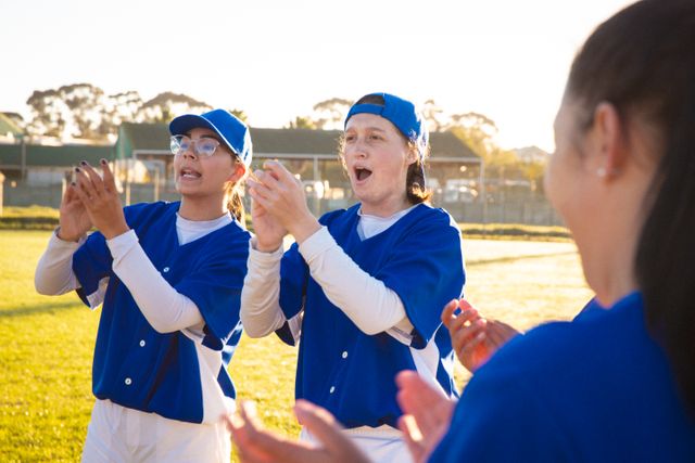 Diverse group of female baseball players in blue uniforms cheering and clapping during a game. Ideal for use in sports-related content, team-building promotions, youth sports programs, and advertisements focusing on diversity and teamwork.