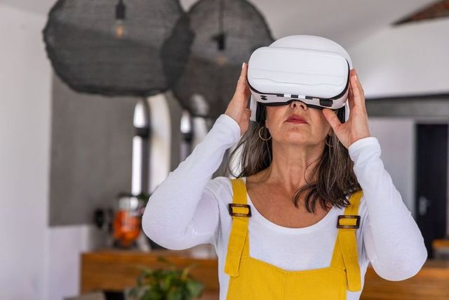 Mature woman exploring virtual reality wearing white headset and yellow overalls in modern kitchen