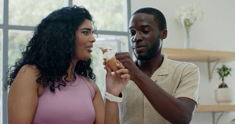Couple sharing melting ice cream cone in sunlit kitchen, playful summer moment