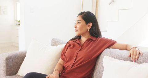 Woman relaxing on light-gray sofa in bright contemporary living room, looking thoughtful