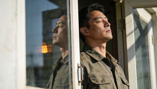Asian man standing in doorway tilting face into sunlight, glass reflection, olive jacket