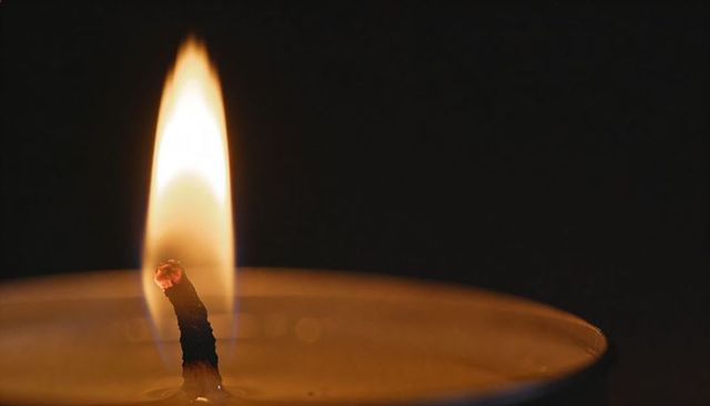 Macro candle wick burning with blue-based yellow flame over molten wax and glowing ember
