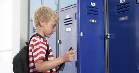 Elementary school boy opening locker holding tablet in sunlit corridor