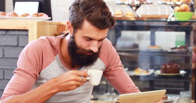 Man enjoying coffee while using tablet in a cozy cafe setting. Ideal for use in lifestyle, technology, or food-related content. Emphasizes relaxing moments, casual dining, and modern habits.