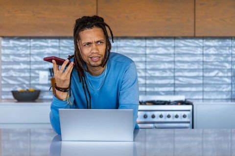 African american man multitasking in modern kitchen, taking call while working on laptop