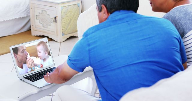 Grandparents are sitting on couch and chatting with their grandchild on a laptop. The smile on their faces shows their happiness and love. They are engaging with their grandchild through technology, bridging the distance between generations. This image is perfect for illustrating concepts of family connections, the elderly engaging with technology, and long-distance relationships.