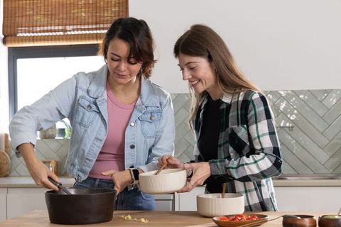 Two diverse women cooking dinner together in sunlit kitchen stirring pot and mixing bowl